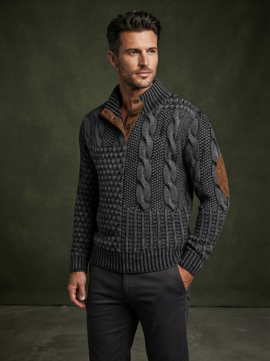 A man wearing a dark gray and black cable-knit zip-up sweater with brown leather elbow patches, styled with dark trousers against a dark olive green studio backdrop.