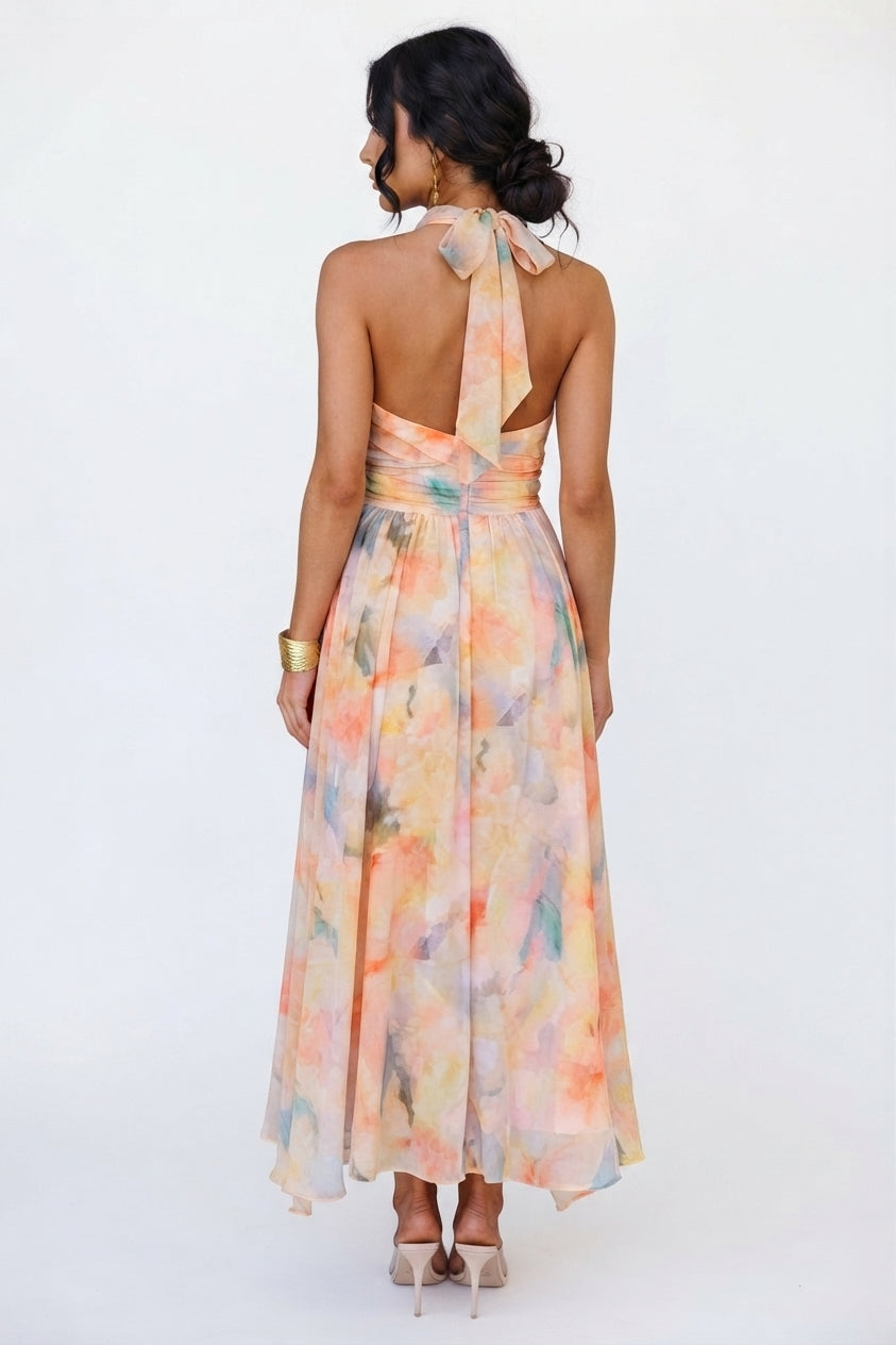 A woman viewed from the back wearing a halter-neck maxi dress with a watercolor floral print in pastel peach, orange, pink, and light blue. The dress features a tie-back at the neck and a flowing skirt, paired with nude high heels against a plain white studio background.