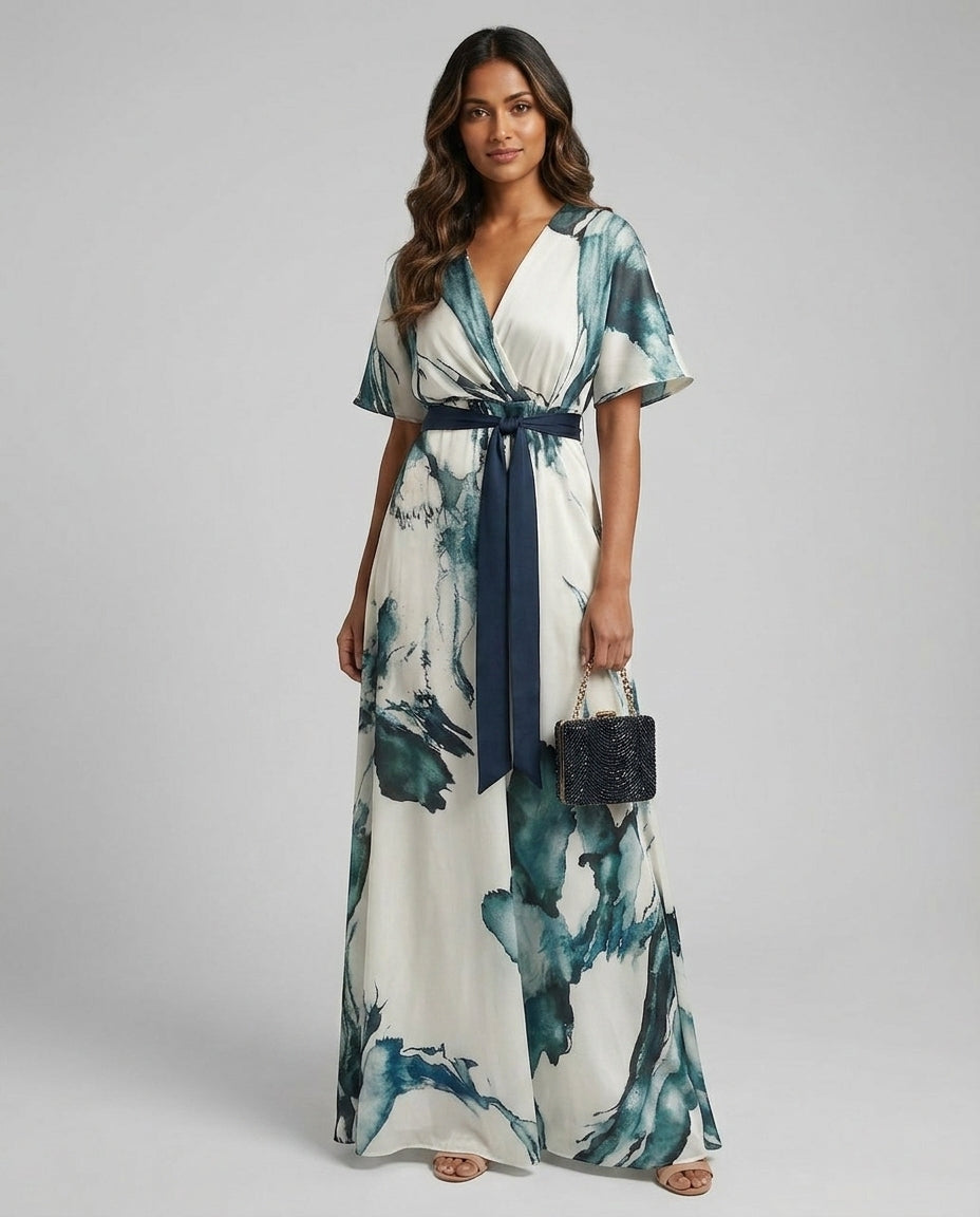 A woman modeling a white maxi dress with a blue and green watercolor-style print, featuring a navy blue tie belt at the waist and holding a small black handbag, set against a plain light gray studio background.