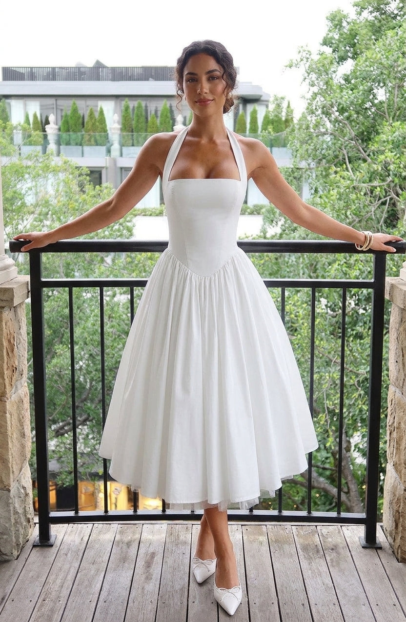 A woman stands on a wooden balcony wearing a white halter-neck midi dress with a structured bodice and full skirt, paired with white pointed heels. Trees and a building are visible in the background behind a black metal railing.