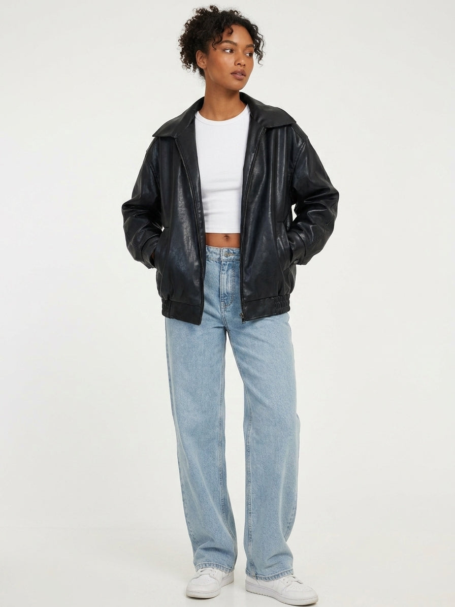 A model wearing a black faux-leather zip-up jacket over a white crop top and light-wash blue jeans, standing against a plain white studio background.
