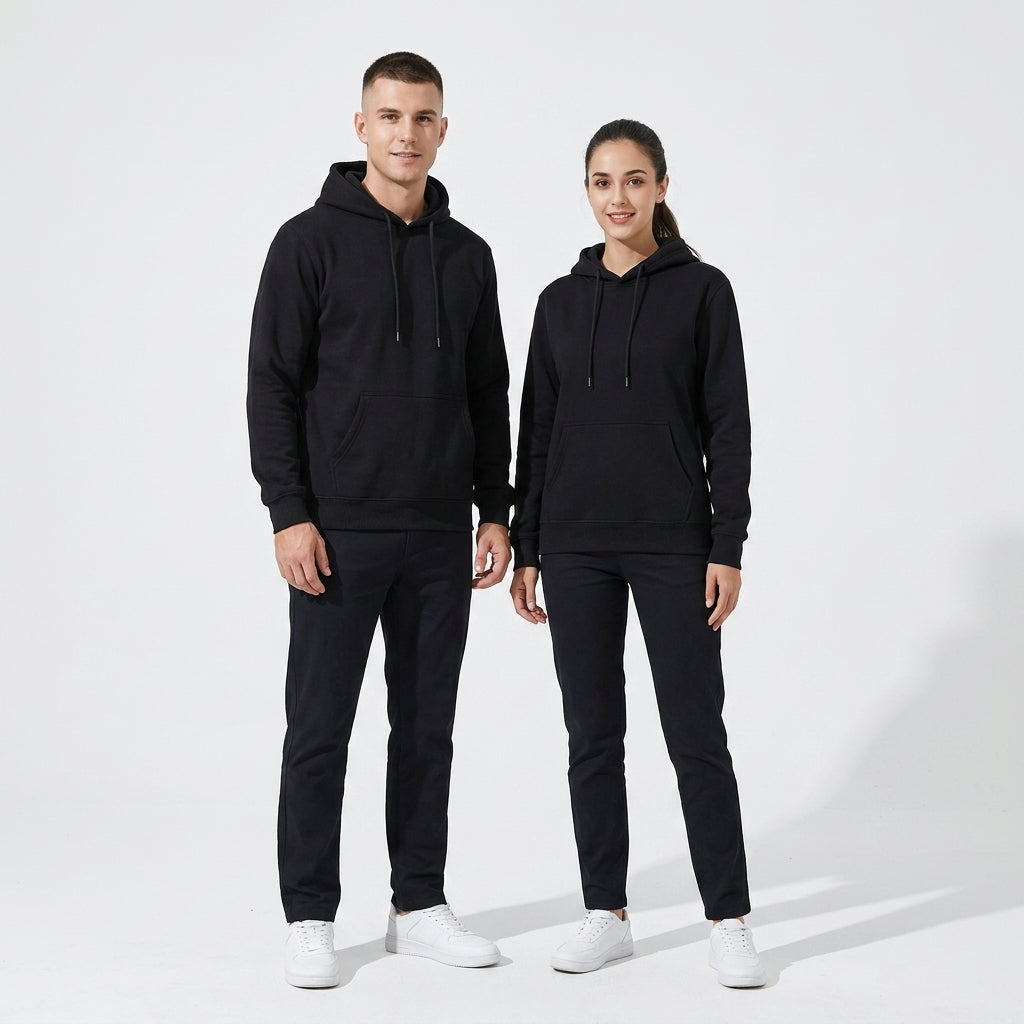 A man and a woman standing side by side wearing matching black hoodies and black joggers, with white sneakers, against a clean white studio background.
