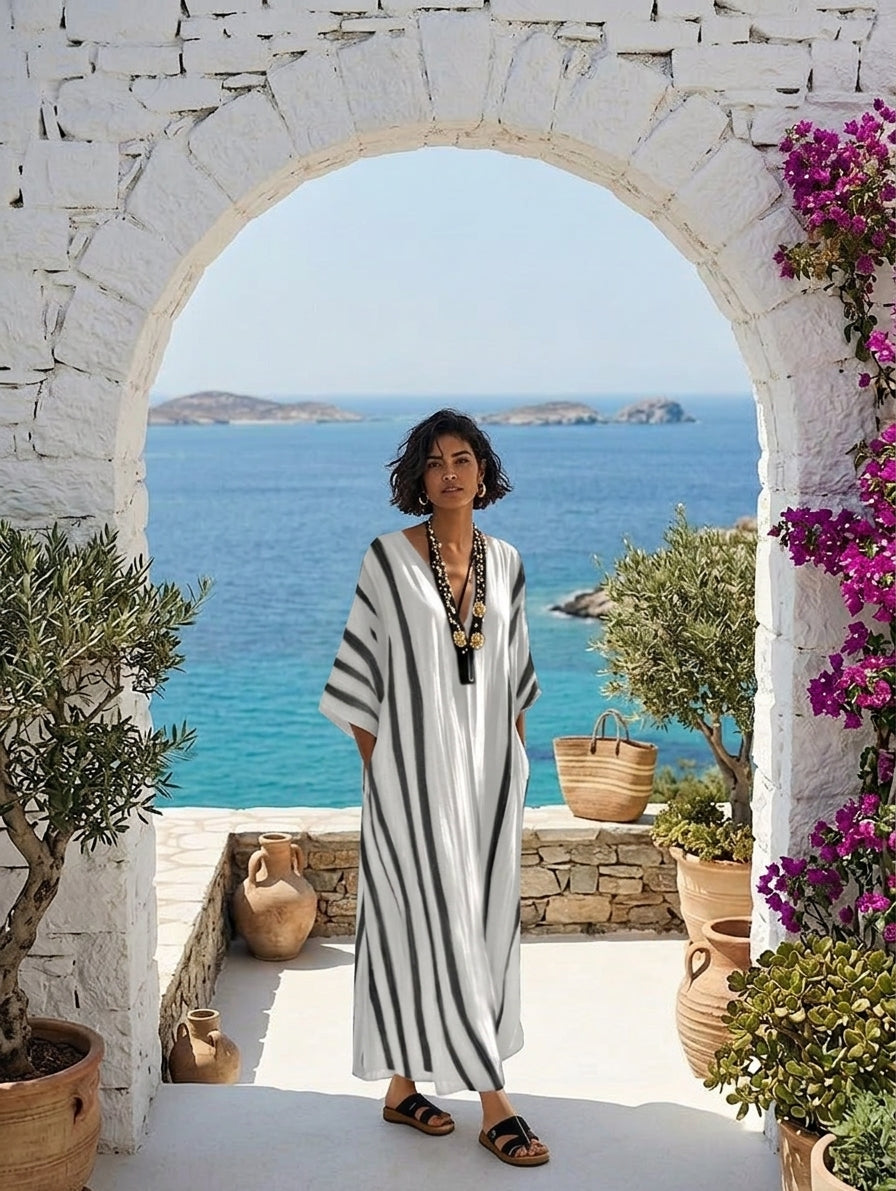 A woman wearing a black and white vertically striped kaftan dress stands framed by a white stone archway, with a scenic blue sea and islands in the background.