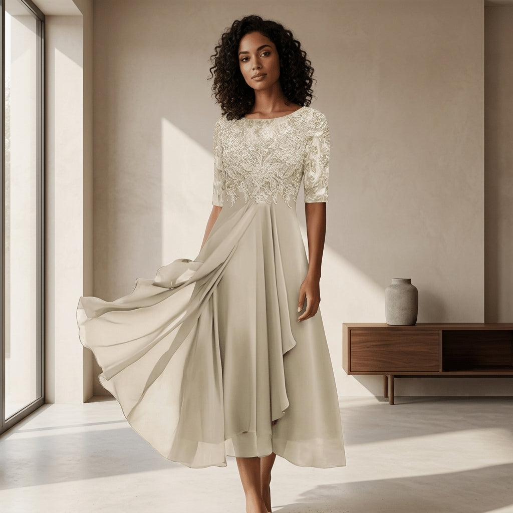 A woman stands indoors wearing a flowing midi dress with a lace bodice and chiffon skirt, posed in a softly lit room with large windows and a wooden side table.