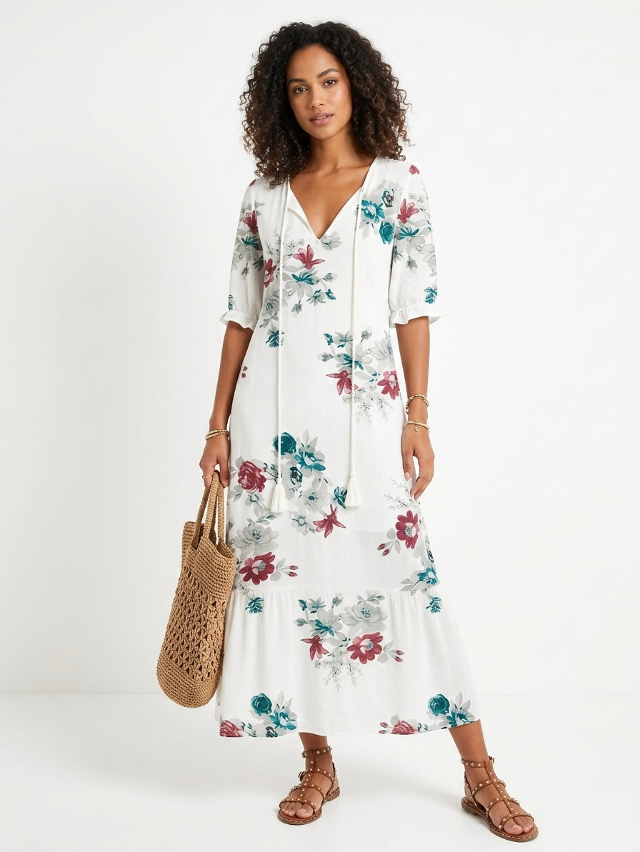 A woman modeling a white midi dress with a colorful floral print, featuring a drawstring V-neckline and elbow-length sleeves. She is holding a woven straw tote bag and wearing brown strappy sandals against a plain white studio background.