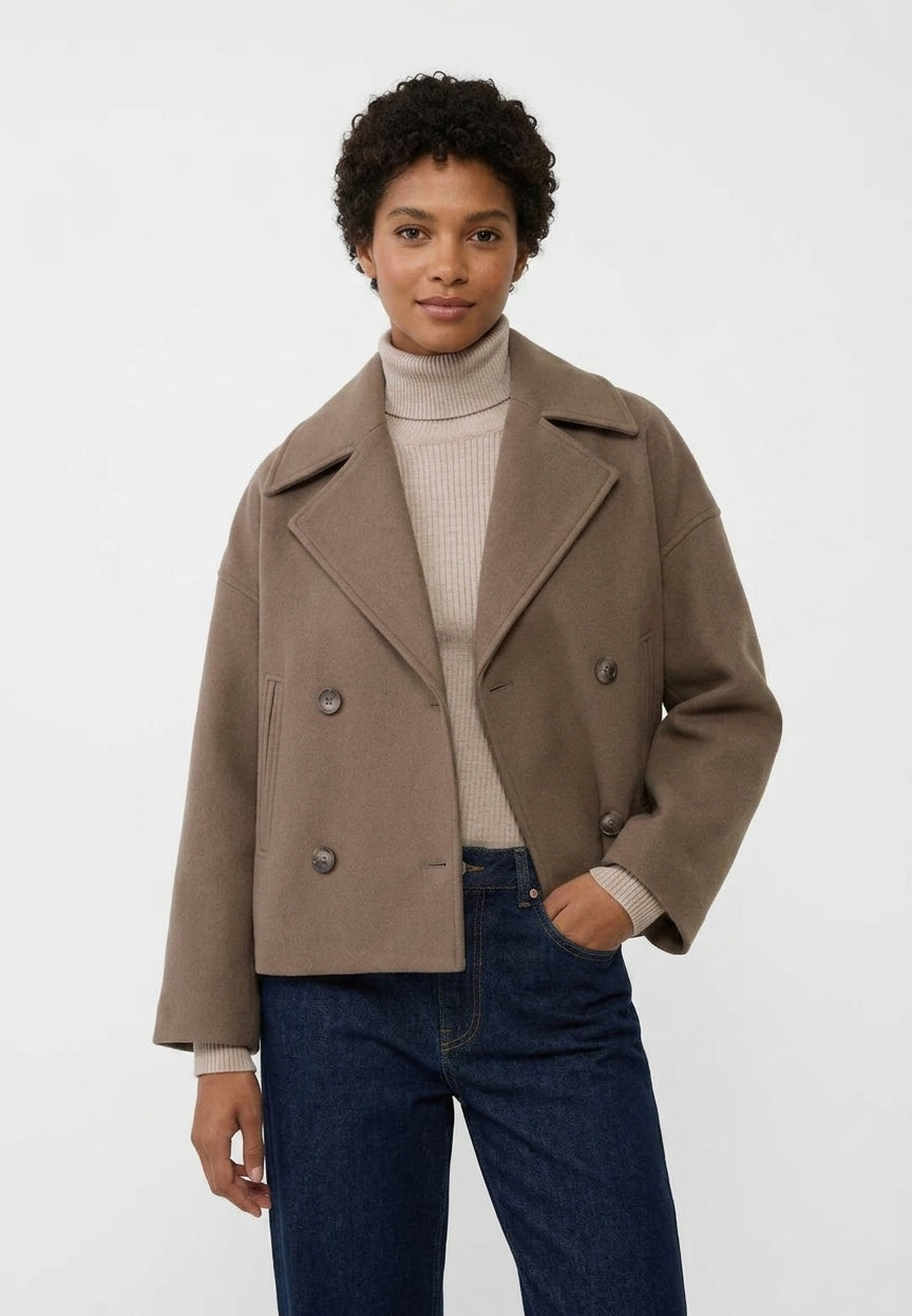 A model wearing a cropped, double-breasted taupe-brown jacket over a beige ribbed turtleneck sweater and dark blue jeans, photographed against a plain light gray studio background.