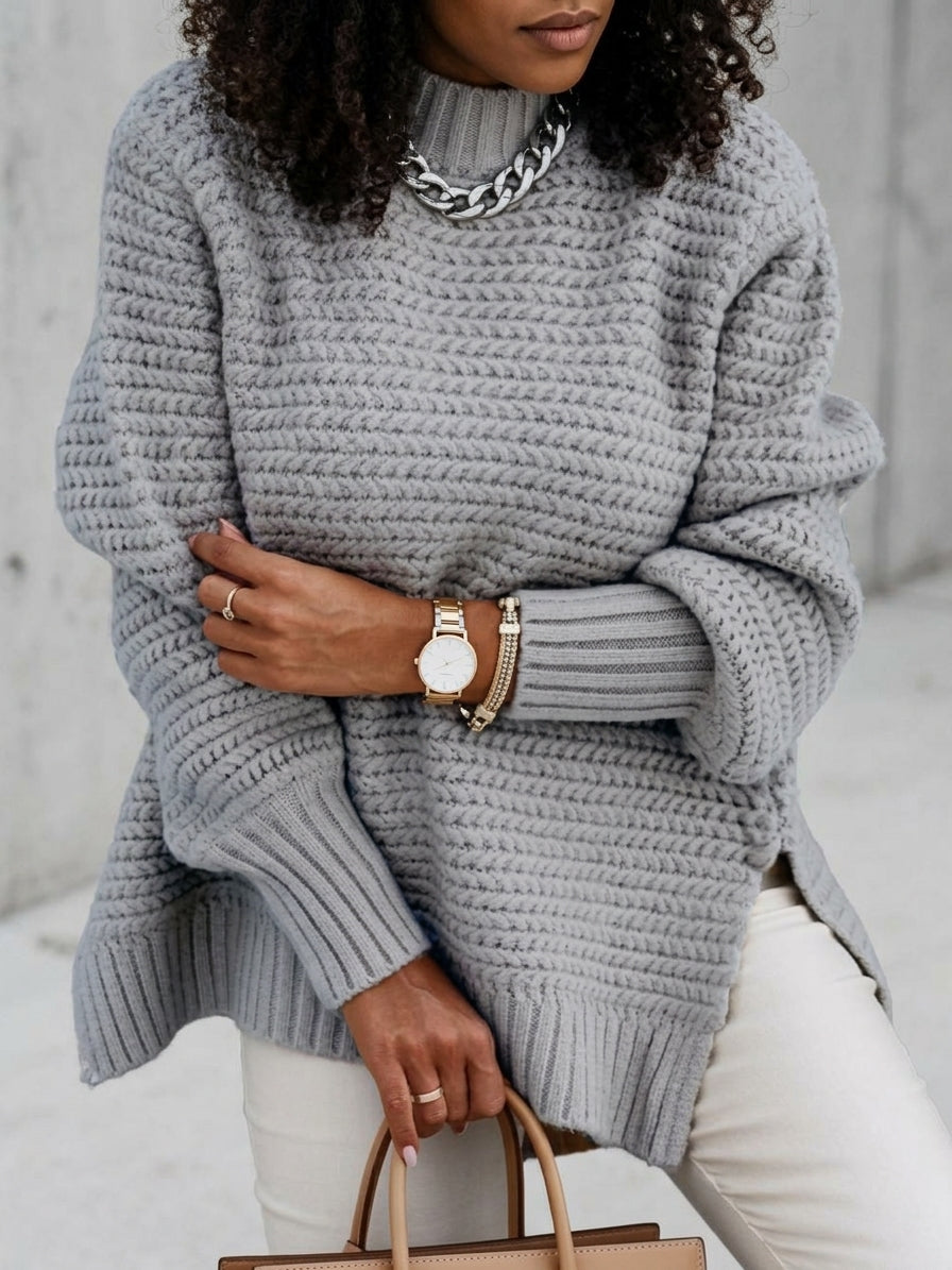 A woman wearing an oversized chunky knit gray sweater, styled with white pants, a chunky silver chain necklace, a gold watch, and bracelets, holding a tan handbag.