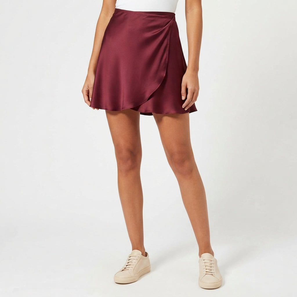 A woman wearing a satin-like wrap mini skirt in burgundy, paired with a white top and beige sneakers, standing against a light gray studio background.