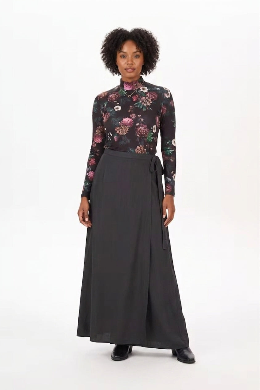 A woman modeling a long-sleeve floral mock-neck top paired with a dark gray wrap-style maxi skirt, standing against a plain white studio background.