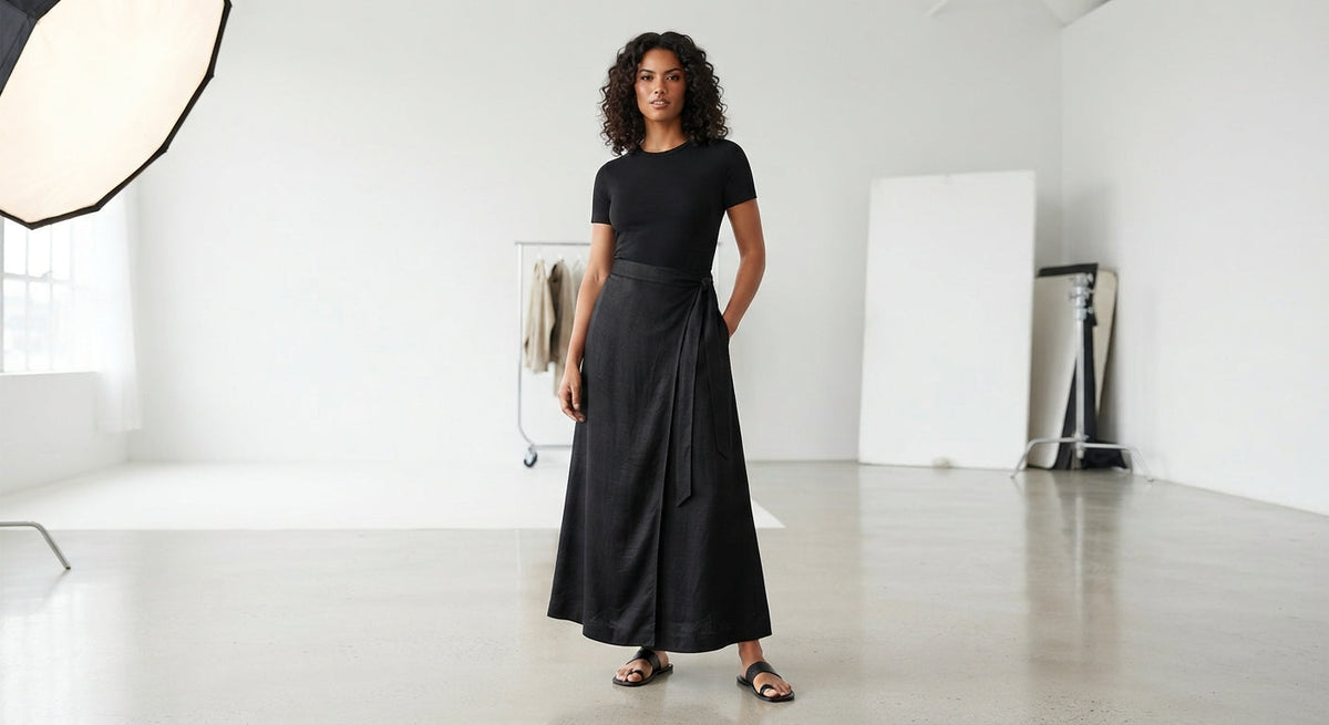 A model wearing a white long-sleeve button-up shirt and a long black skirt, paired with beige loafers, against a plain white studio background.