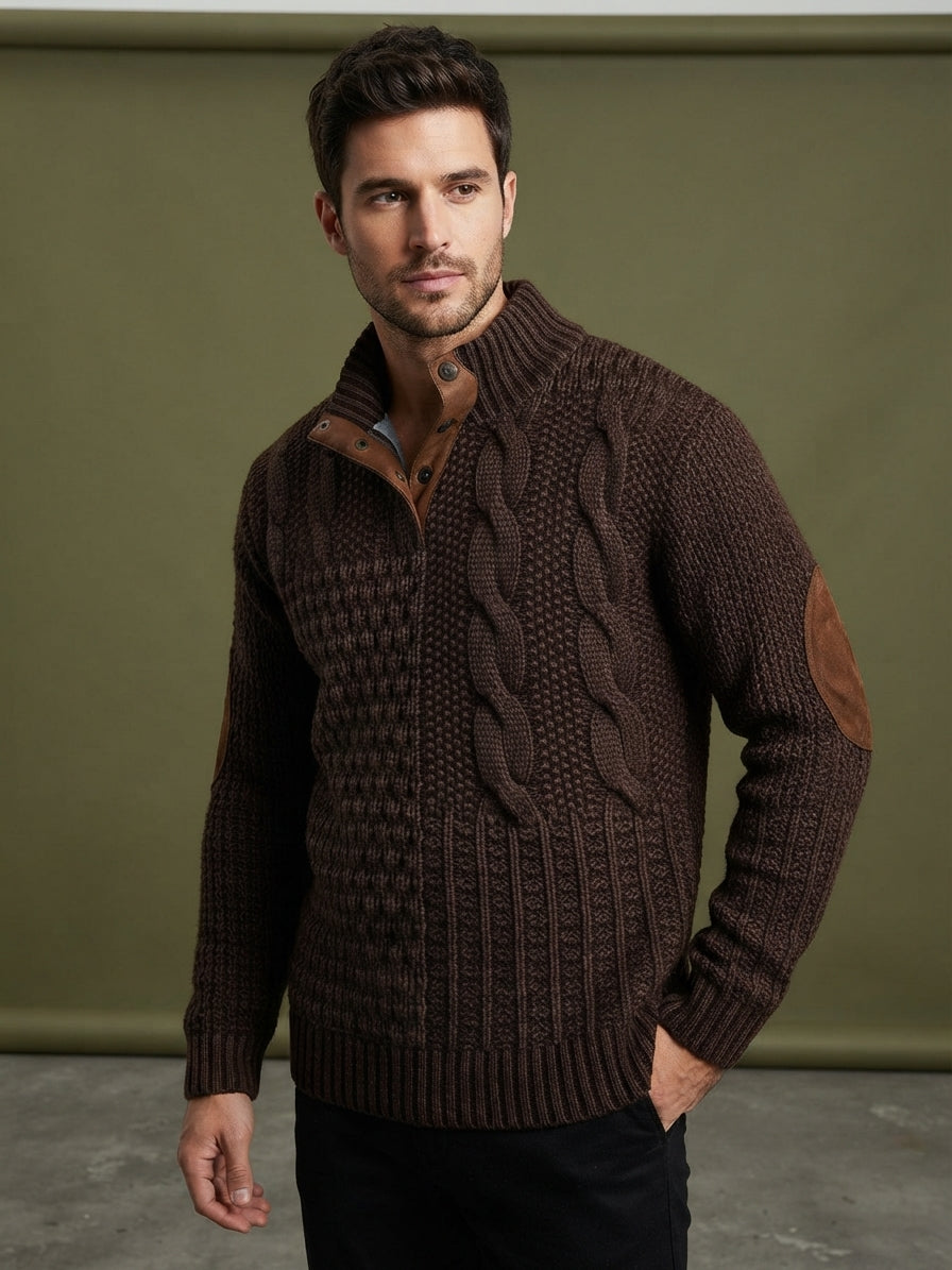 A man wearing a dark brown cable-knit zip-up sweater with brown leather elbow patches, posed against a muted olive green studio backdrop.