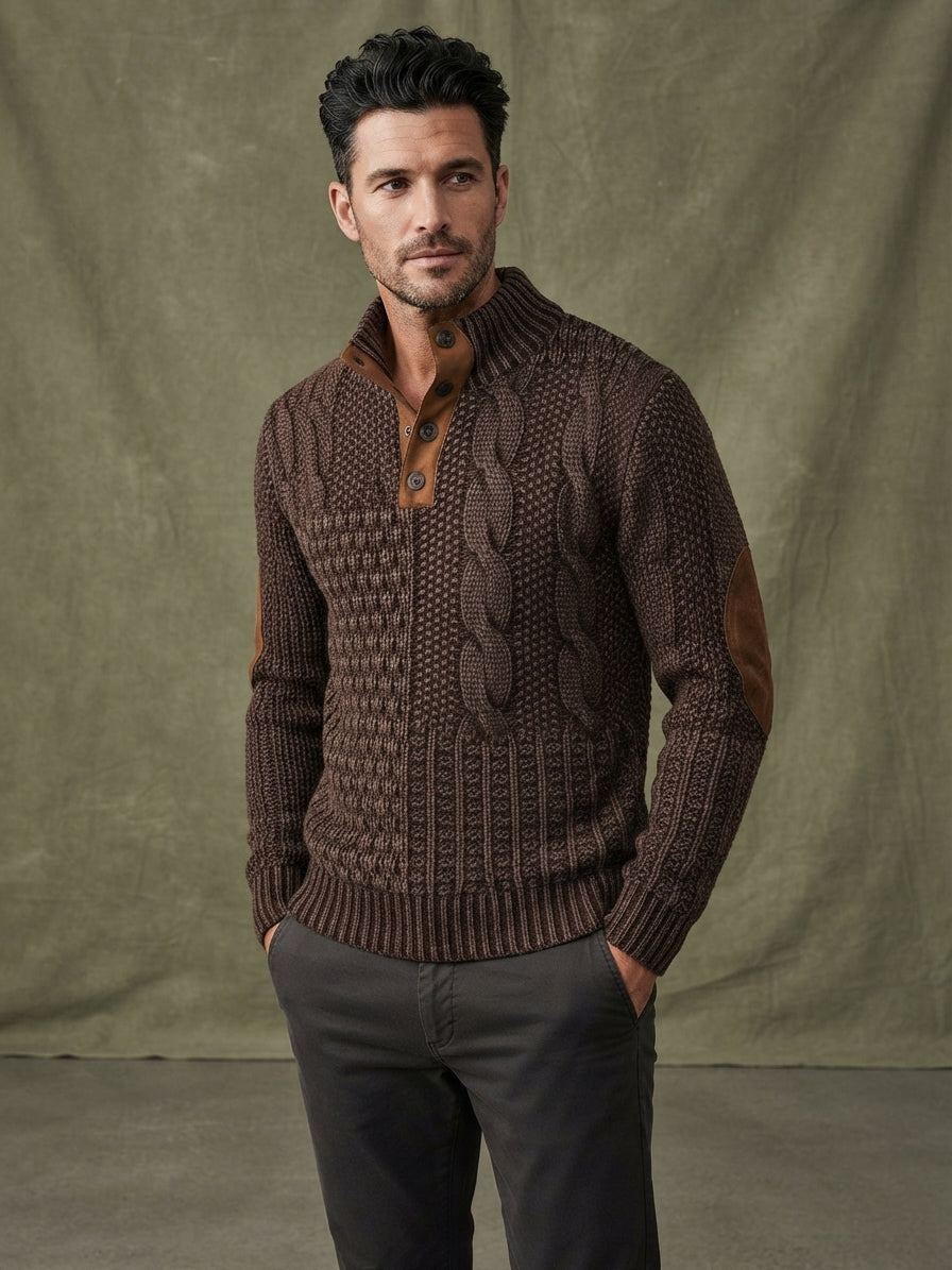 A man wearing a brown cable-knit sweater with brown leather elbow patches and a buttoned collar, paired with dark gray trousers, standing against a textured olive green backdrop.