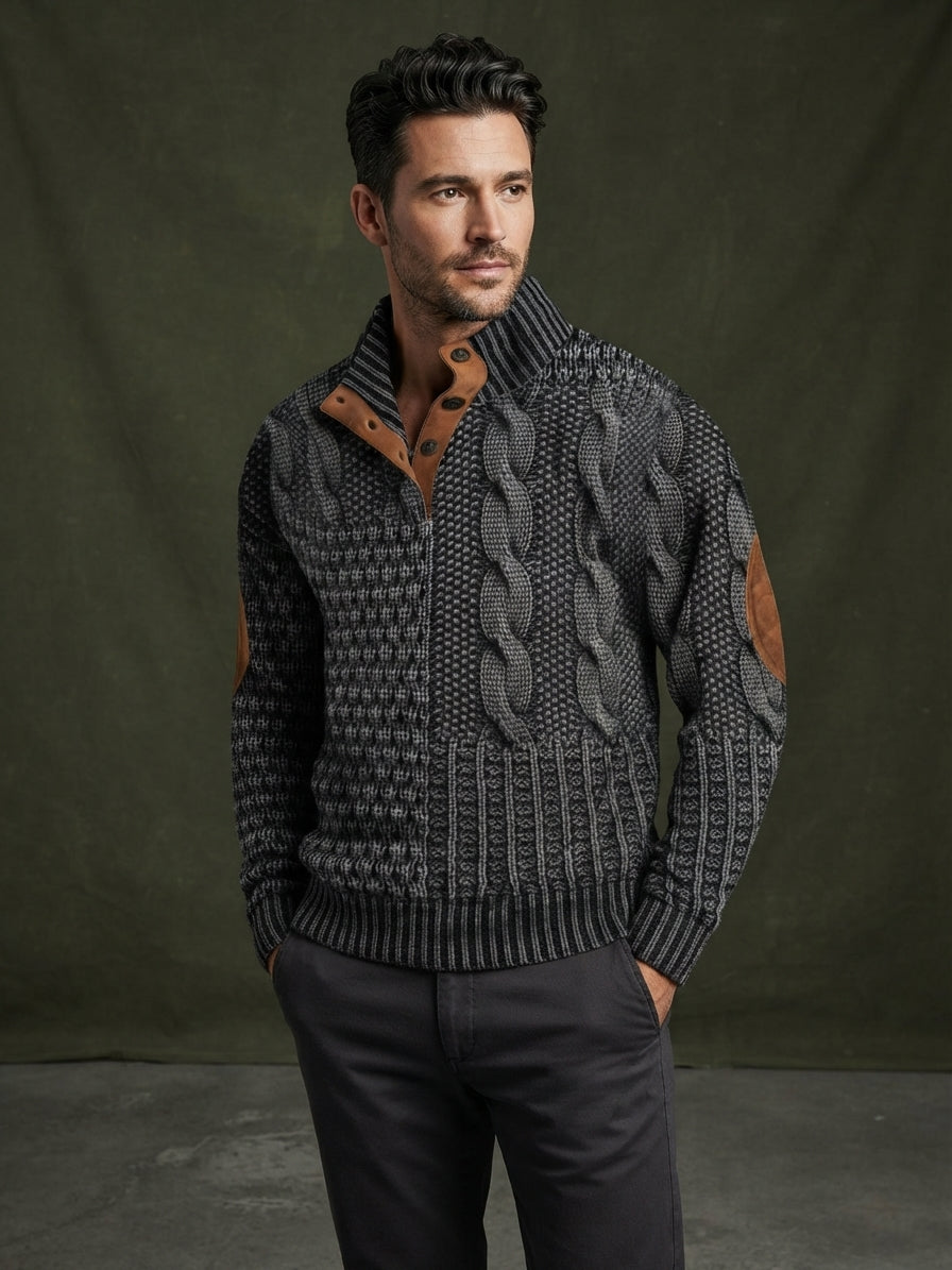 A man wearing a dark gray and black cable-knit zip-up sweater with brown leather patch details on the chest and sleeves, paired with dark gray trousers, set against a dark olive green studio backdrop.