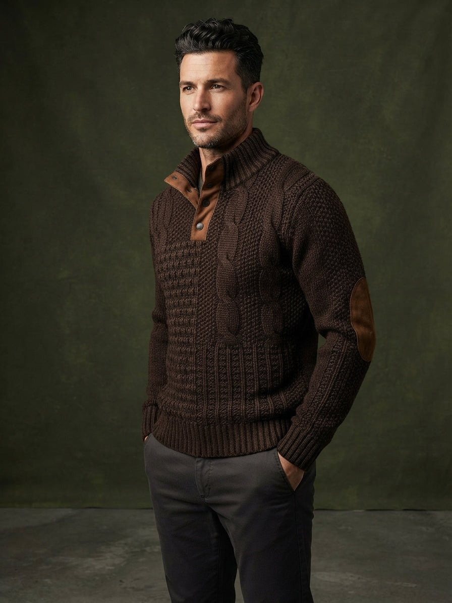 A man wearing a dark brown textured knit sweater with a mock neck, brown button placket, and brown elbow patches, paired with dark gray trousers, posed against a dark olive green studio backdrop.