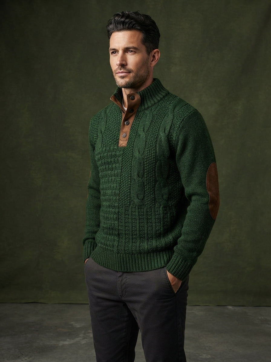 A man wearing a dark green cable-knit sweater with brown elbow patches and a brown button placket, paired with dark gray trousers, standing against a dark olive green studio backdrop.