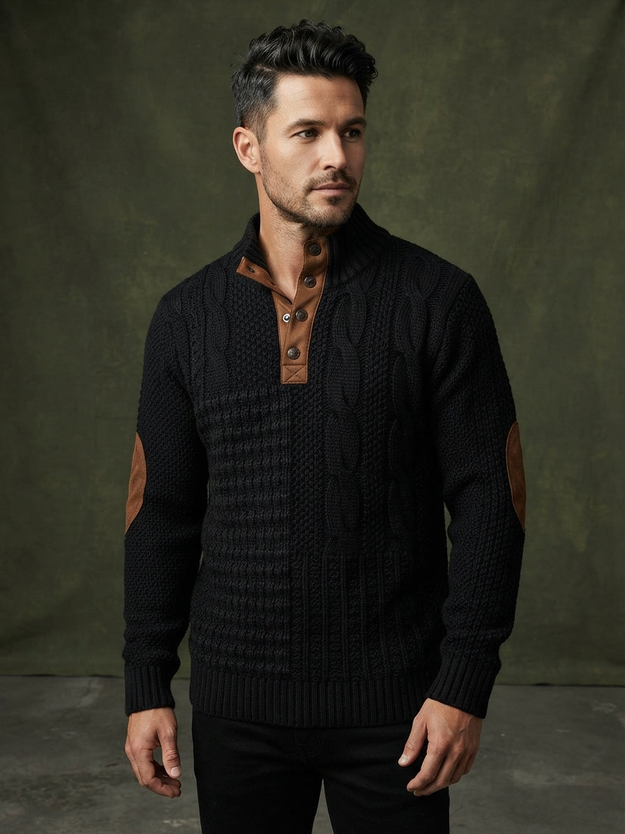 A man wearing a black cable-knit sweater with brown elbow patches and a brown buttoned placket, standing against a dark olive-green studio backdrop.