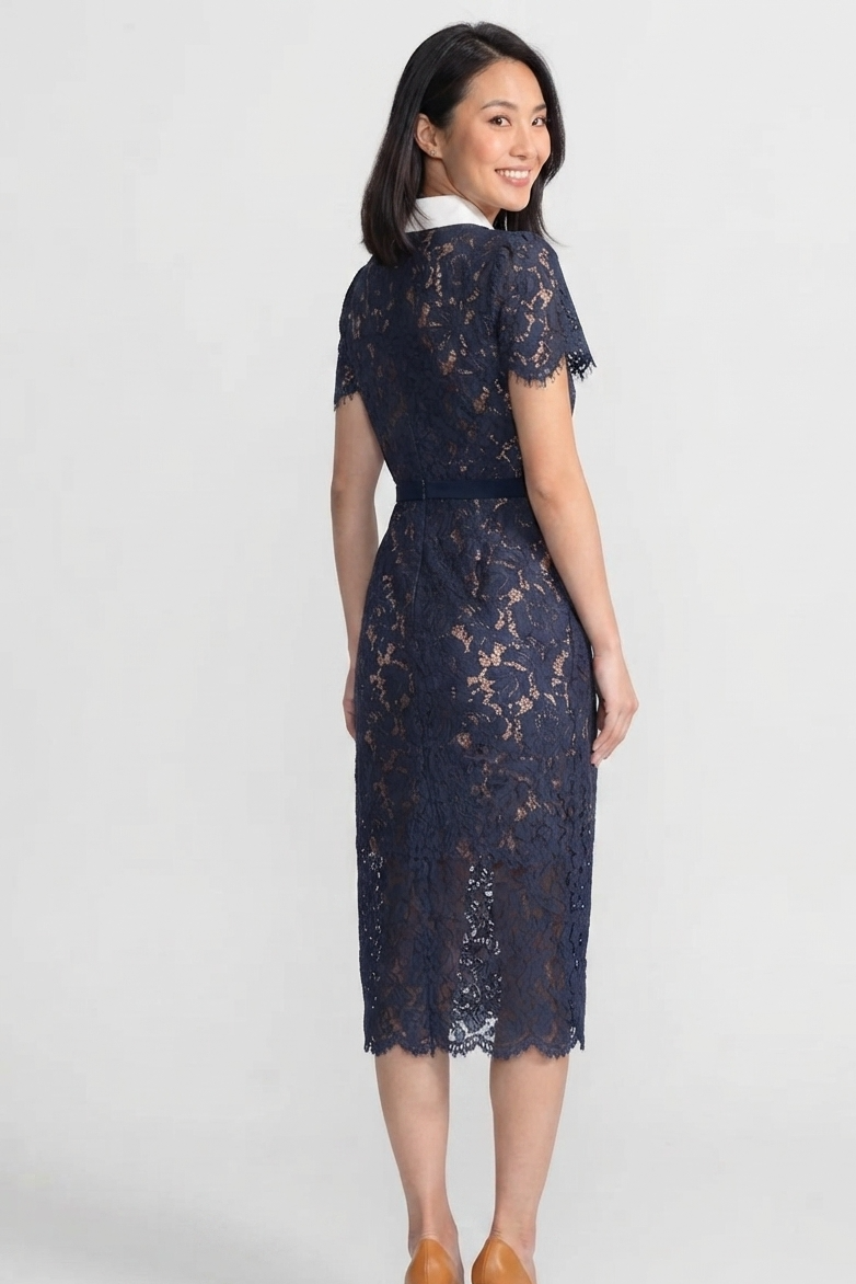 A woman modeling a navy blue lace midi dress with short sleeves, a white collar, and a matching navy belt, shown from a three-quarters back view against a light gray studio background.