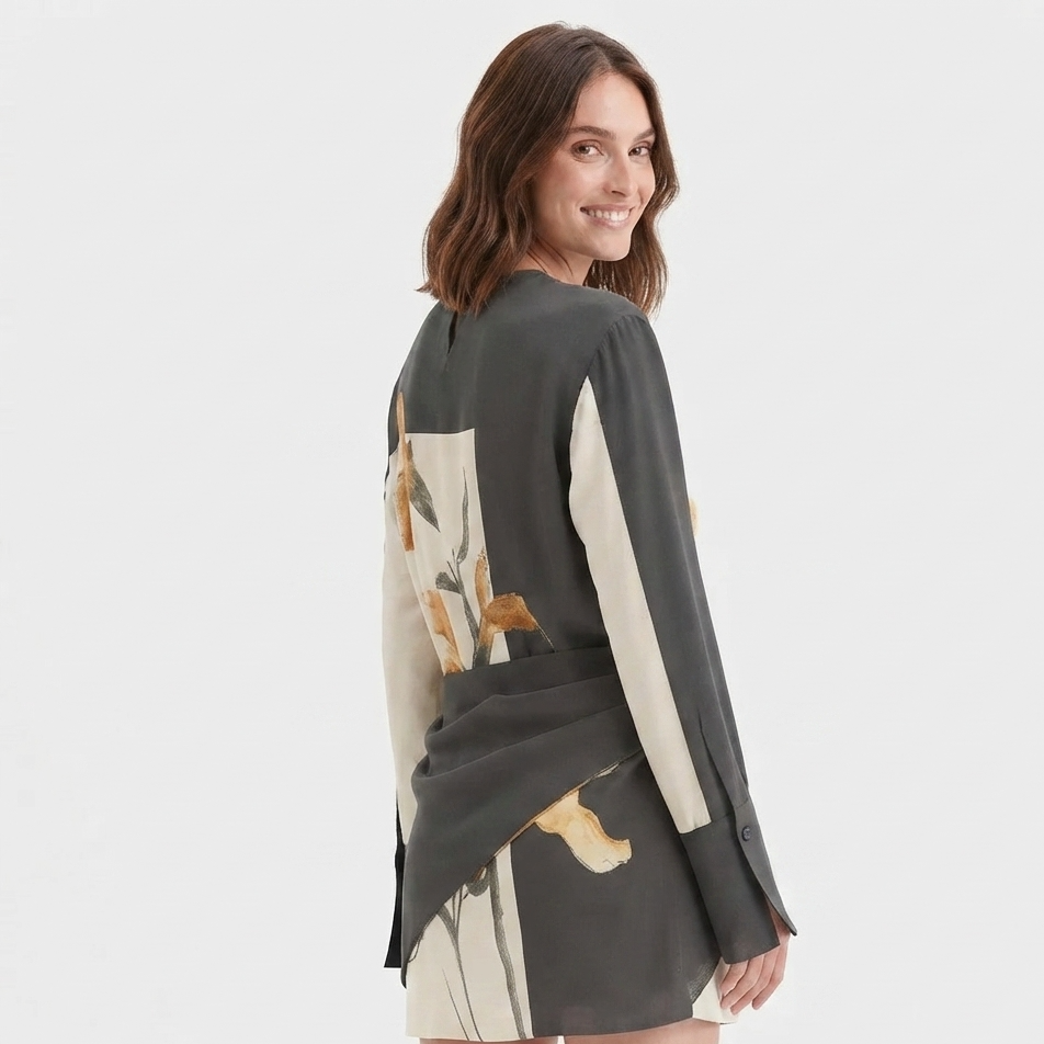 A woman modeling a long-sleeve wrap-style top with a dark gray base and beige panels featuring a botanical print, shown from a three-quarters back view against a light gray studio background.