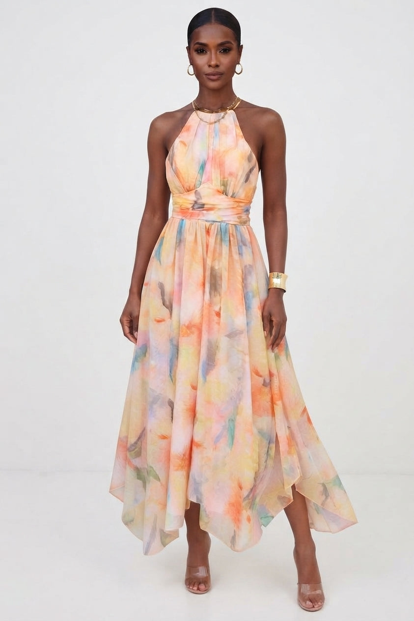 A woman modeling a sleeveless halter-neck maxi dress with a soft watercolor floral print in pastel orange, yellow, pink, and light blue on a white studio background.