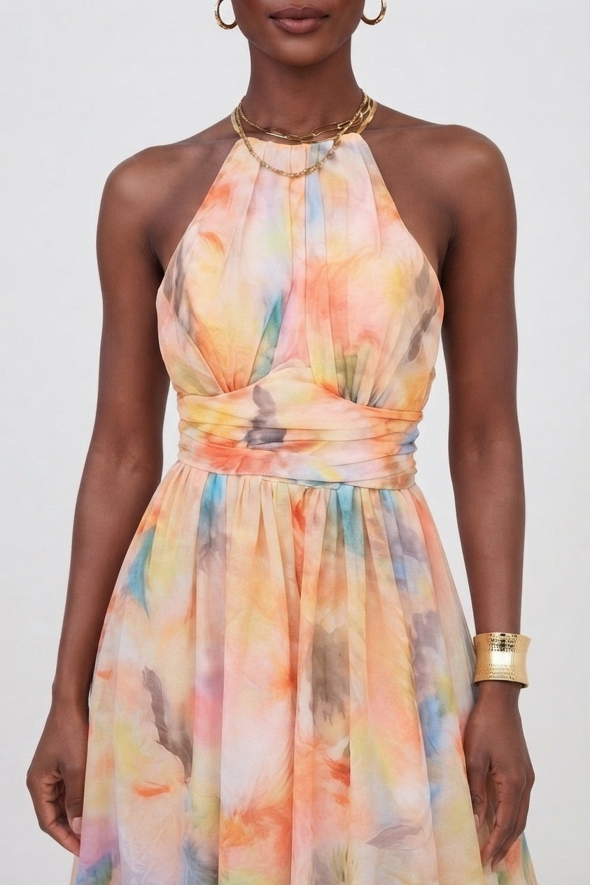 A woman wearing a sleeveless halter-neck dress with a soft watercolor floral print in pastel orange, yellow, pink, blue, and beige tones. She is accessorized with gold jewelry against a plain light gray studio background.