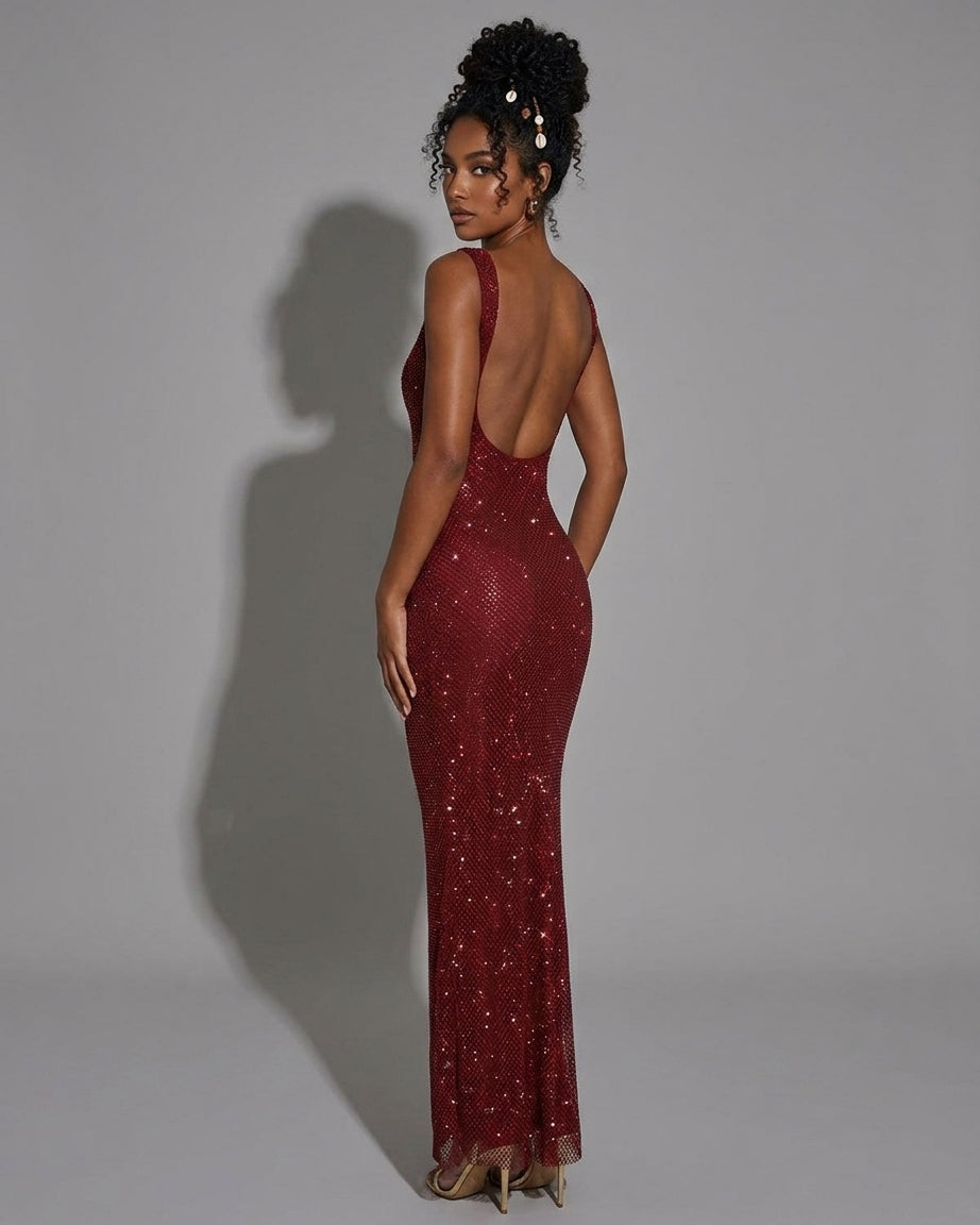 A woman modeling a floor-length, open-back burgundy evening gown covered in sparkling sequins, shown from the back against a plain gray studio background.
