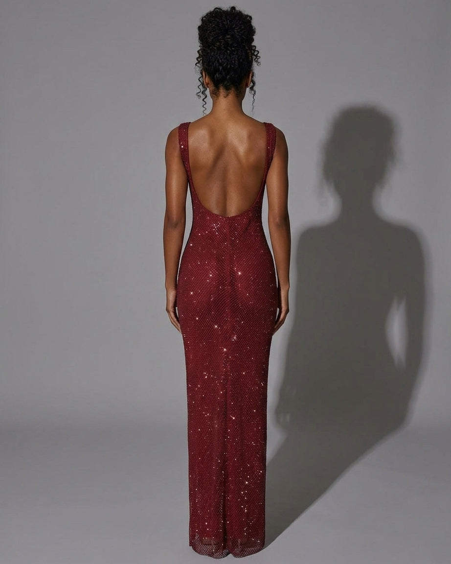 A woman viewed from the back wearing a sleeveless, open-back floor-length gown covered in sparkling sequins. The dress is a deep burgundy red, and the model stands against a plain gray studio background with a prominent shadow cast to the right.