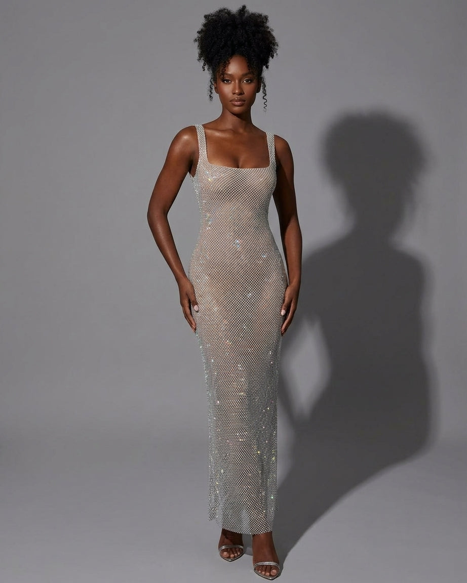 A woman modeling a sleeveless, floor-length sheer mesh gown covered in shimmering sequins, standing against a plain gray studio background with a strong shadow cast to the right.