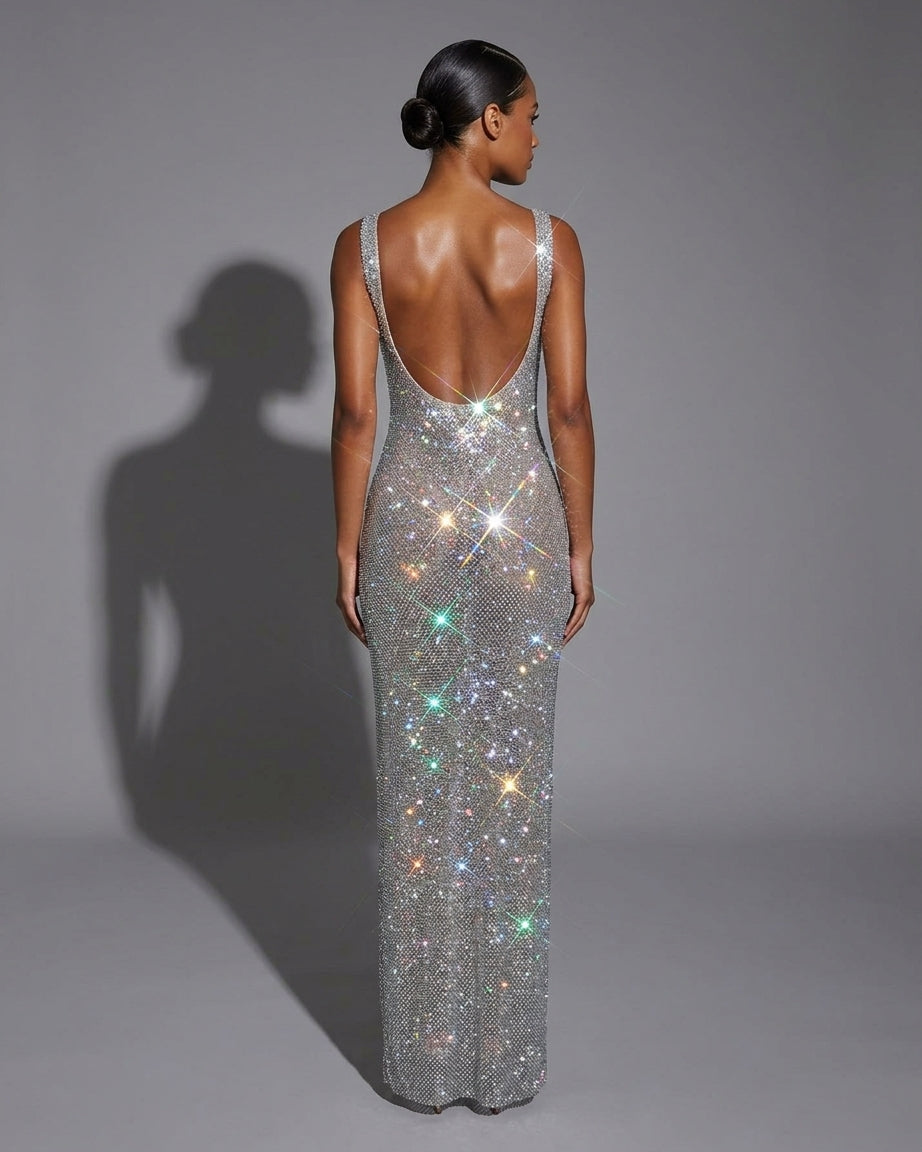 A woman stands with her back to the camera wearing a floor-length, open-back evening gown covered in sparkling silver rhinestones that catch the light with rainbow reflections, against a plain gray studio background.