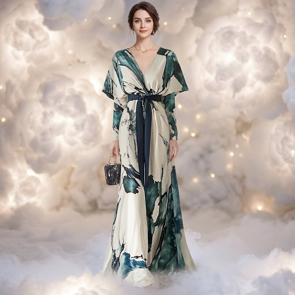 A woman stands in a cloud-like studio setting wearing a long wrap-style dress with a blue and green abstract print on an off-white base, holding a small dark handbag.