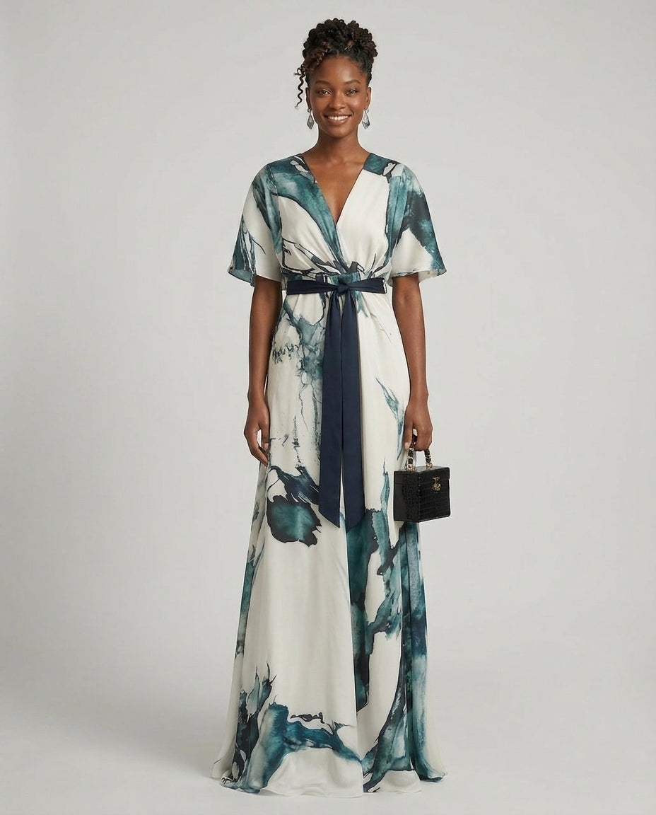 A woman modeling a white maxi dress with a blue and green abstract watercolor print, featuring a V-neckline, short flutter sleeves, a matching dark blue tie belt, and holding a small black handbag against a light gray studio background.