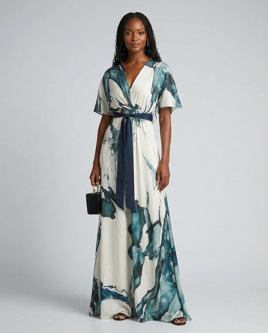 A woman modeling a long white maxi dress with a blue and teal abstract watercolor print, featuring a V-neckline, short flutter sleeves, a matching navy blue waist tie belt, and holding a small black handbag against a light gray studio background.