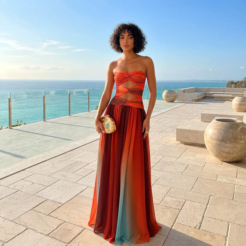 A woman stands on a sunlit stone terrace overlooking the ocean, wearing a strapless maxi dress with a bright orange-red bodice and a flowing gradient skirt that transitions through red, orange, and teal. She holds a small patterned clutch.