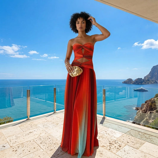 A woman stands on a sunlit terrace overlooking a turquoise ocean and rocky coastline, wearing a strapless red-orange gown with a cutout waist and a long flowing skirt. She holds a metallic gold clutch.