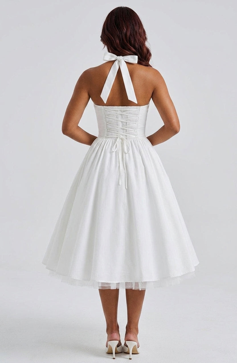 A woman shown from the back wearing a white halter-neck A-line dress with a corset-style bodice and a full skirt, paired with white heels, against a light gray studio background.