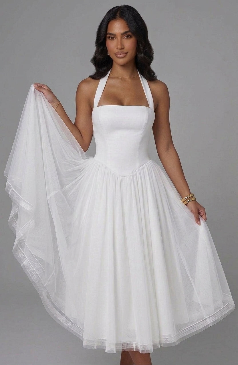 A woman modeling a white halter-neck dress with a fitted bodice and a full, flowing tulle skirt, holding out one side of the sheer skirt to show its drape against a plain gray studio background.