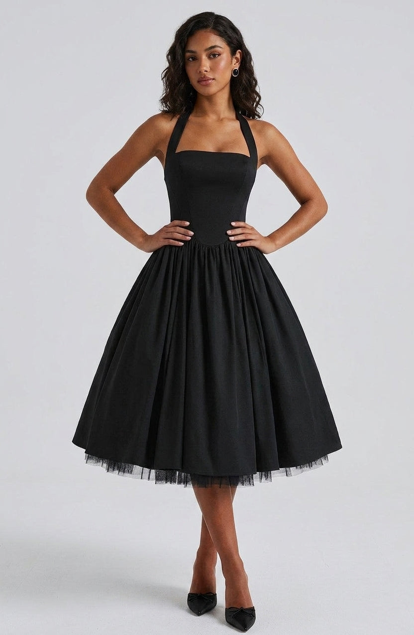 A woman modeling a black halter-neck swing dress with a full skirt and tulle hem, paired with black pointed-toe heels, against a light gray studio background.