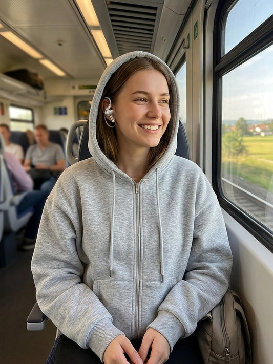 A woman smiling while sitting on a train, wearing a light gray zip-up hoodie. The photo is taken inside the train next to a window, with other passengers blurred in the background.