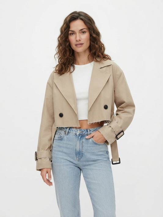 A woman modeling a cropped beige double-breasted blazer with black buttons, worn over a white crop top and light blue jeans, against a plain white studio background.