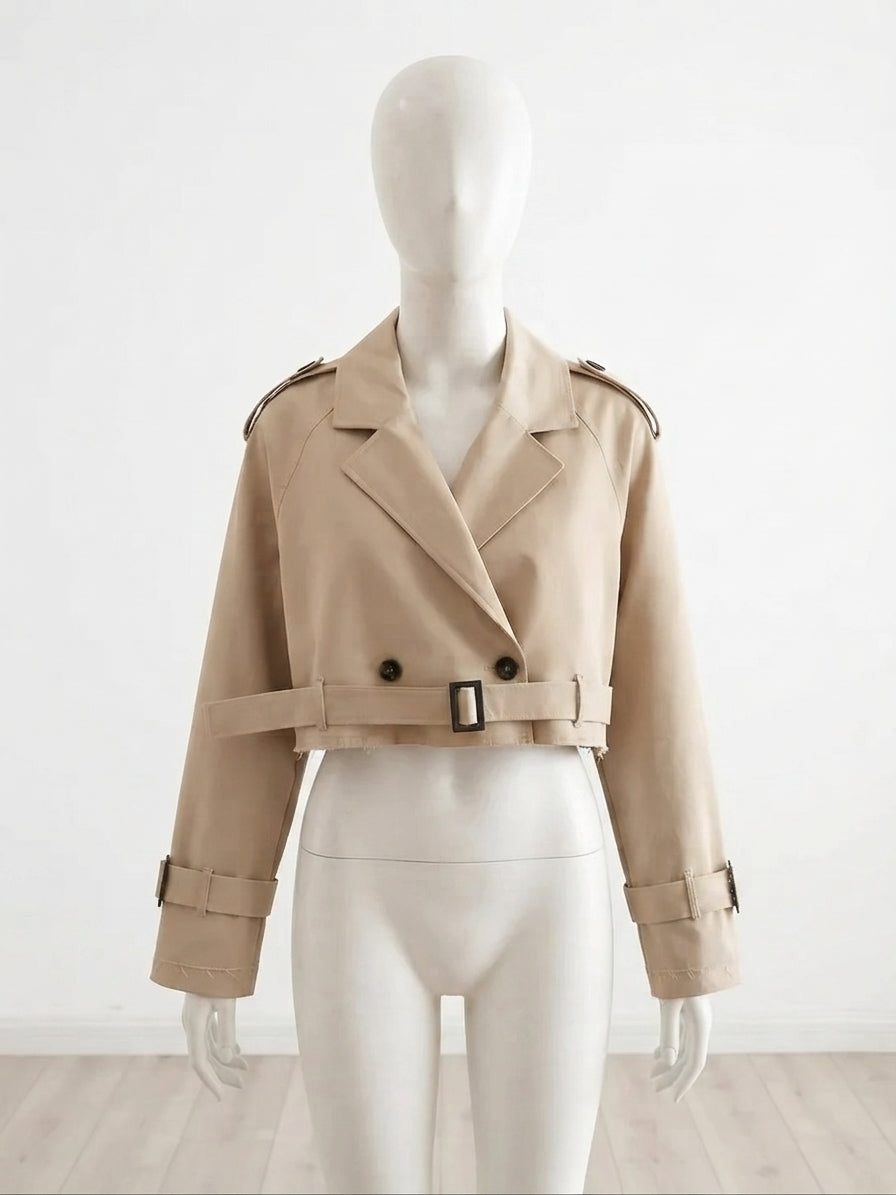 A cropped beige trench jacket displayed on a white mannequin against a plain white wall and light wood floor.