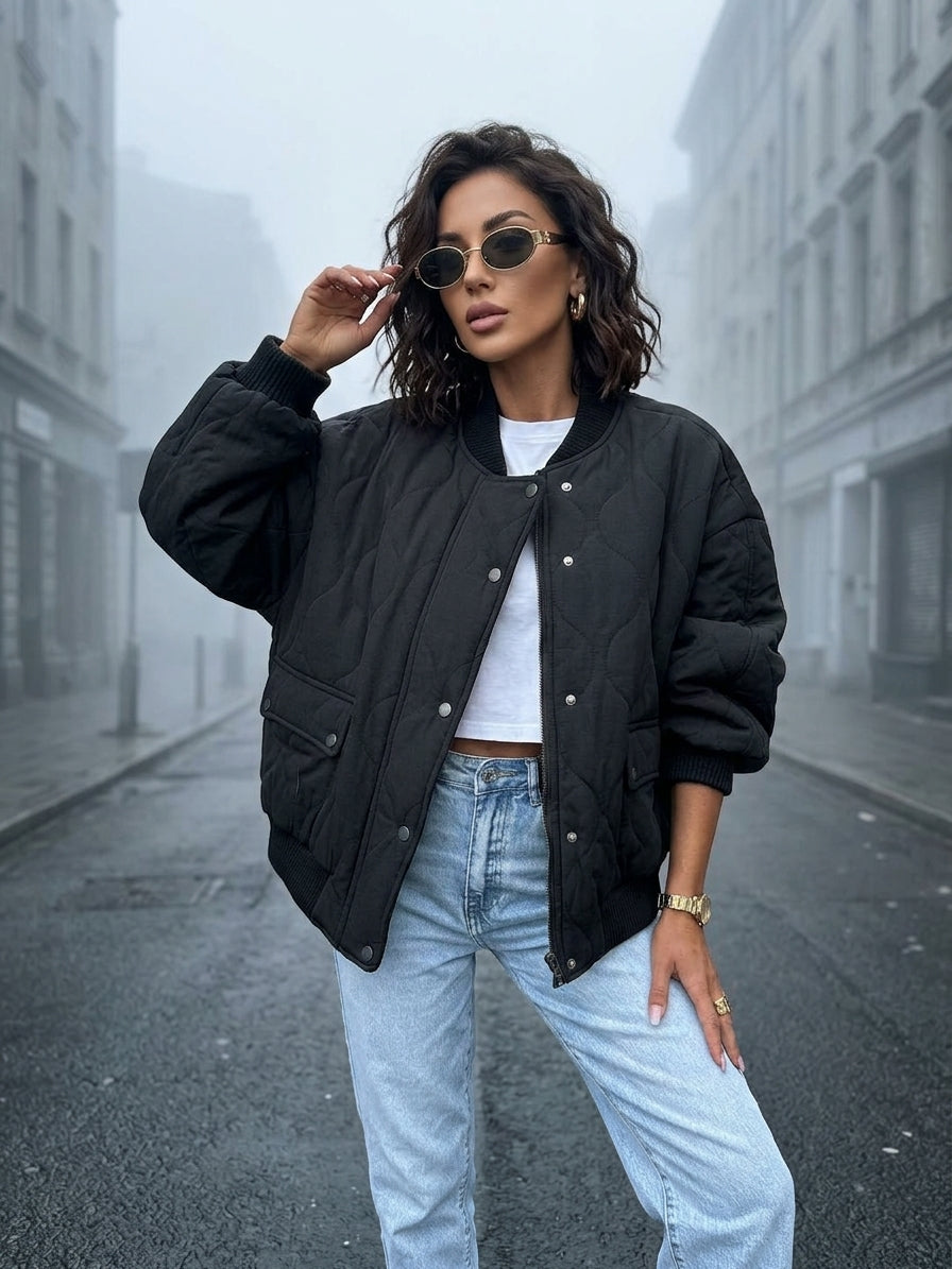 A woman stands on a foggy city street wearing a black quilted bomber jacket over a white cropped T-shirt, light blue jeans, and round sunglasses.