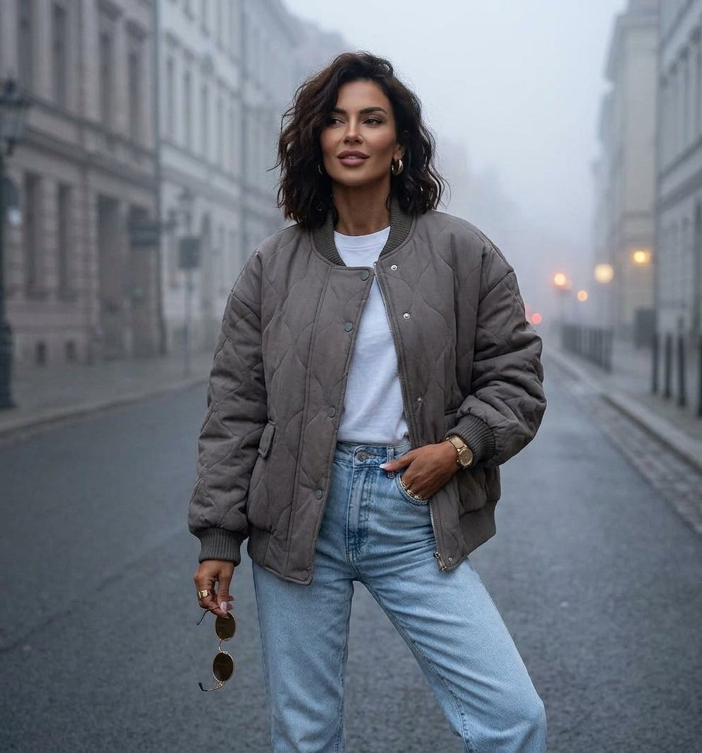 A woman stands on a city street wearing a taupe quilted bomber jacket over a white t-shirt and light blue jeans.
