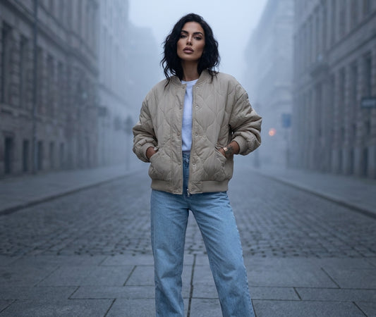 A woman stands on a foggy city street wearing a beige quilted bomber jacket over a white top and light blue jeans.