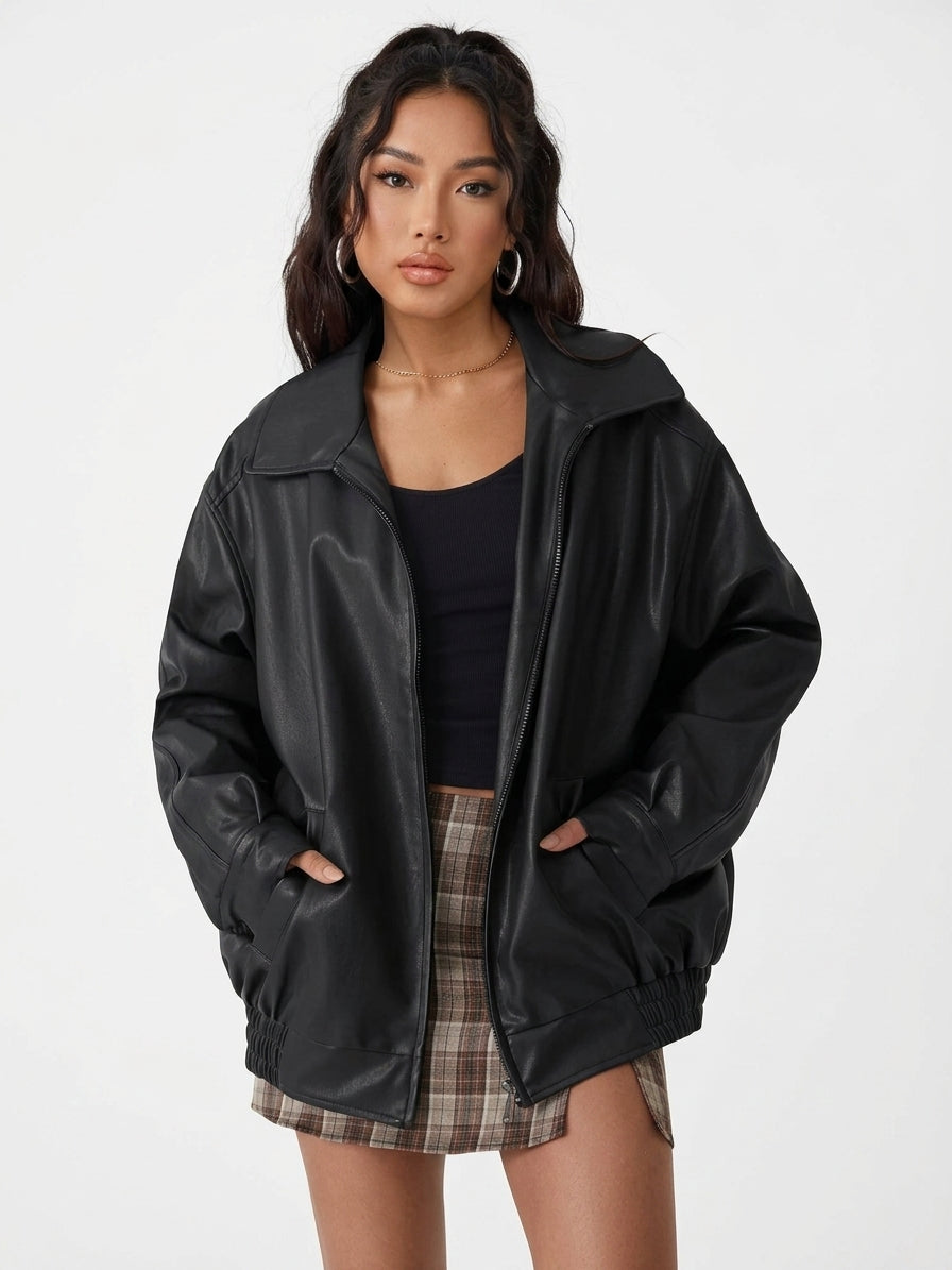 A woman modeling an oversized black faux-leather jacket with a high collar and front zipper, worn over a black top and a brown plaid mini skirt, against a plain light gray studio background.