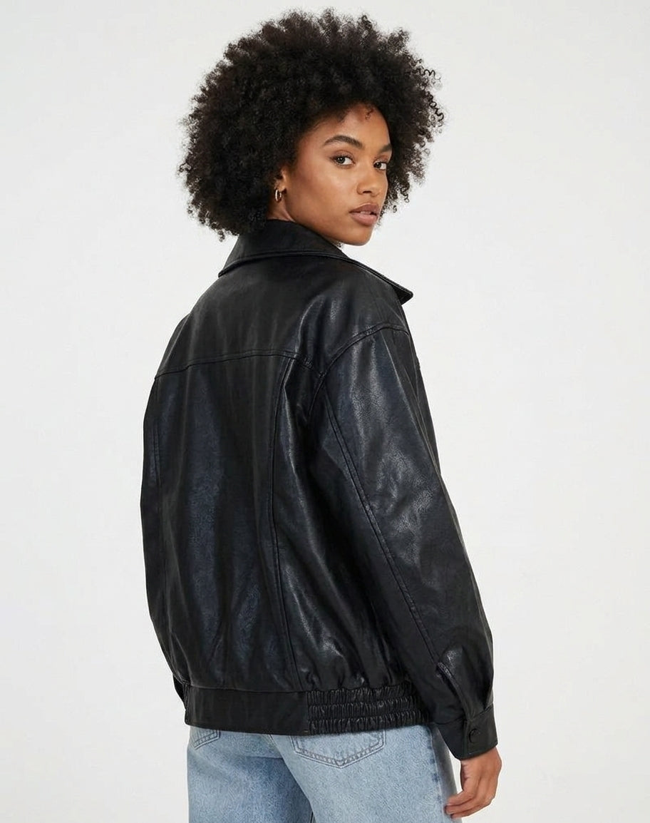 A woman with an afro hairstyle wearing a black faux leather jacket and light blue jeans, photographed from a three-quarters back view against a plain white studio background.
