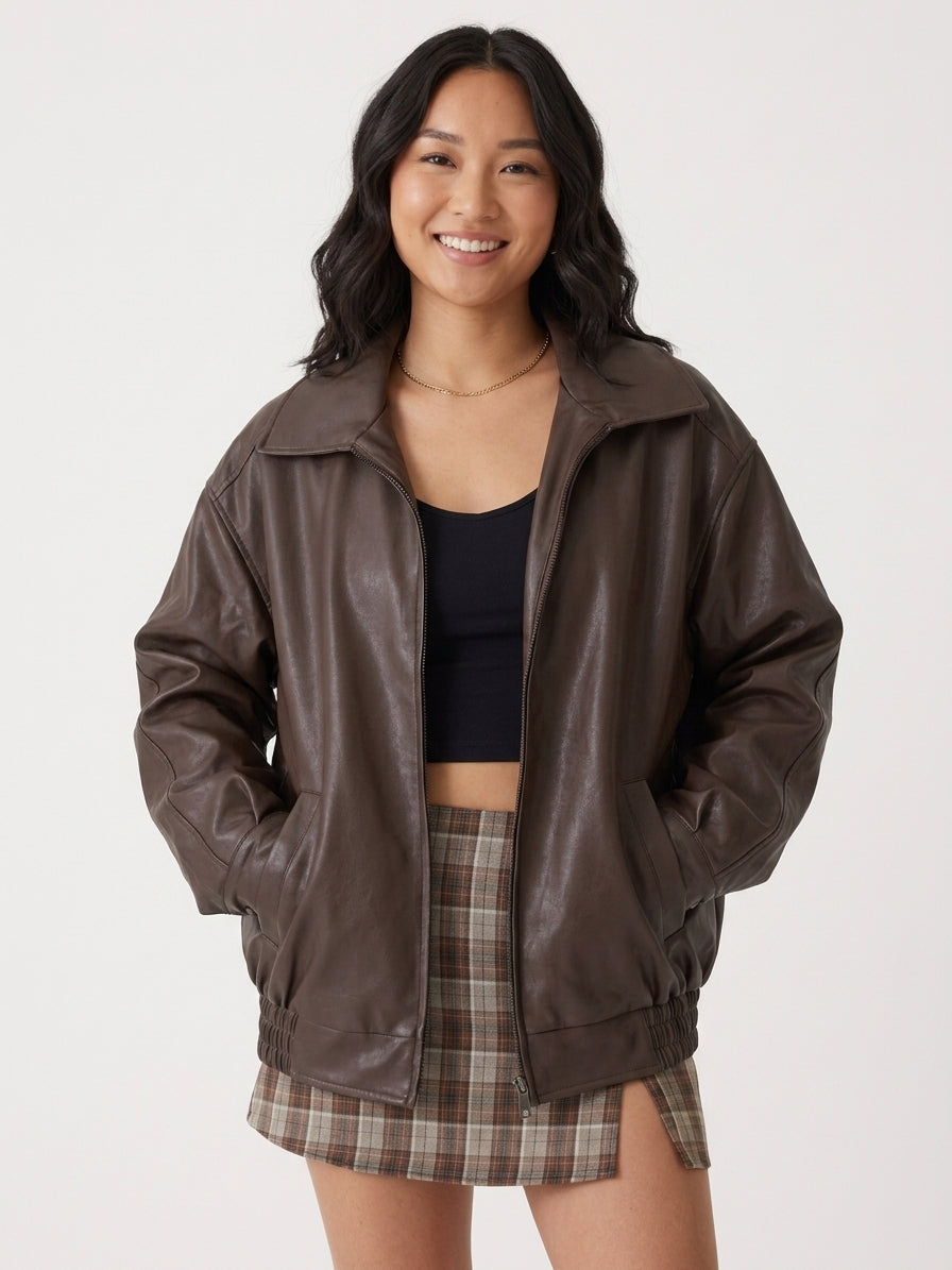 A woman modeling an oversized dark brown faux-leather jacket, styled with a black crop top and a brown plaid mini skirt, against a plain light gray background.