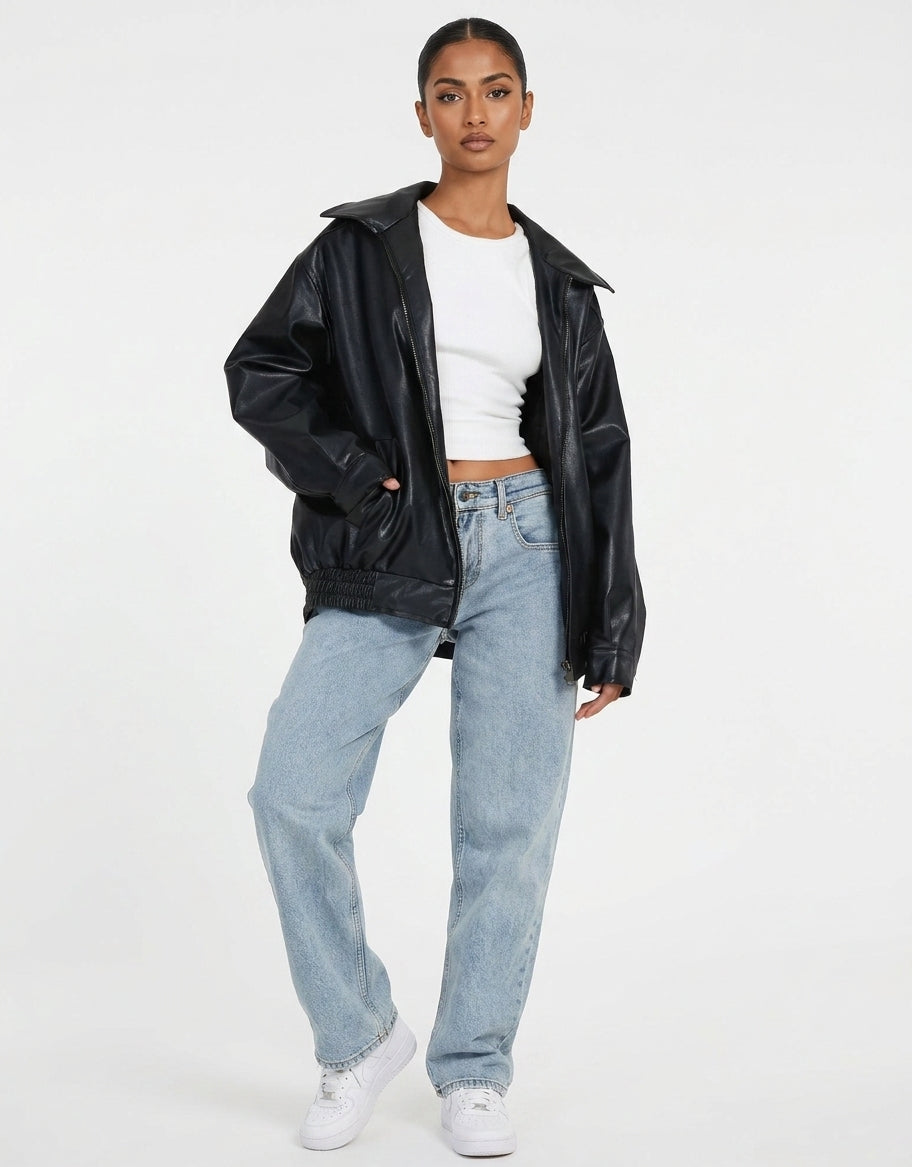 A model wearing a black faux-leather jacket, light-wash blue jeans, a white crop top, and white sneakers, standing against a plain white studio background.