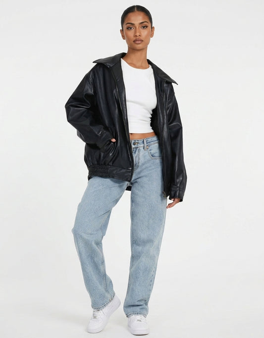 A model wearing a black faux-leather jacket, light-wash blue jeans, a white crop top, and white sneakers, standing against a plain white studio background.