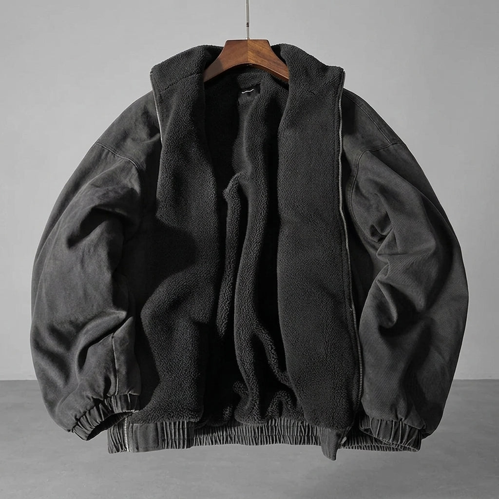A dark gray zip-up jacket with a plush black fleece lining, hanging on a wooden hanger against a neutral gray background.