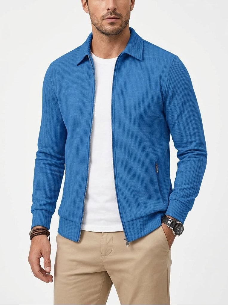 A man wearing a bright blue textured zip-up jacket over a white t-shirt and khaki pants, standing against a light gray studio background.