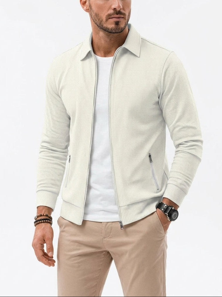 A man wearing a light cream ribbed zip-up jacket over a white t-shirt and beige trousers, standing against a plain white studio background.